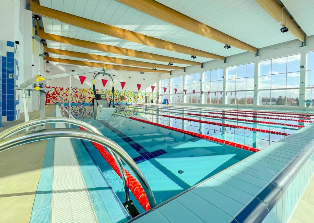 Na zdjęciu widać basen z wyznaczonymi torami, oddzielonymi czerwonymi linami. Po prawej stronie znajdują się duże okna, przez które wpada światło dzienne, rozjaśniając całą halę. Na suficie widać drewniane belki i jasne panele z lampami. Z tyłu, przy końcu niecki, jest kolorowa dekoracja na ścianie i czerwone chorągiewki, które oznaczają koniec toru. Przy brzegu stoją metalowe poręcze, dzięki którym łatwiej wejść do wody. Basen wygląda czysto i zadbanie, widać, że wszystko jest dobrze utrzymane.