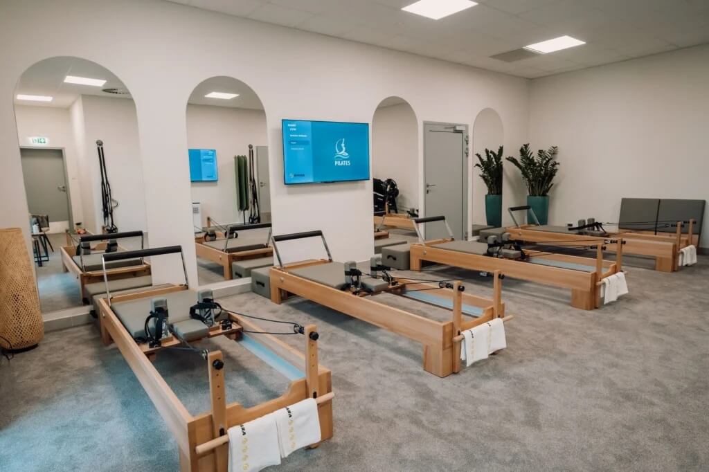 Na zdjęciu widzimy eleganckie, nowocześnie urządzone studio Pilates. W pomieszczeniu znajdują się cztery drewniane reformery — profesjonalne maszyny do ćwiczeń — ustawione w dwóch równych rzędach. Każdy z nich wyposażony jest w pasy, sprężyny i ruchome platformy, a na końcu każdego leży starannie złożony biały ręcznik, co podkreśla dbałość o komfort i higienę.