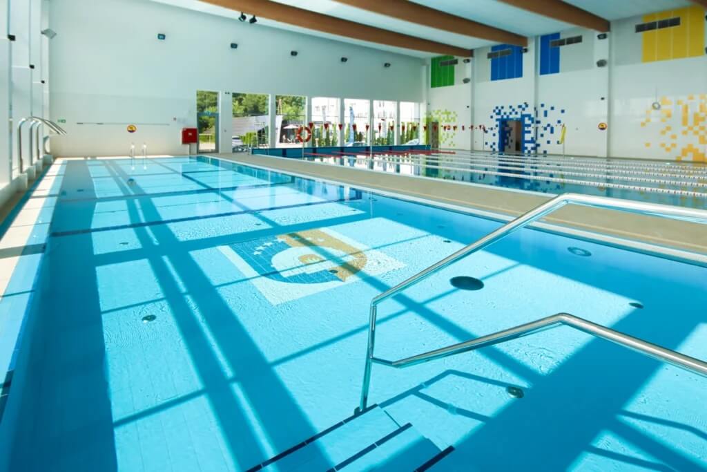 Na zdjęciu widzimy nowoczesną halę basenową o jasnym, przyjaznym charakterze. Woda w basenie jest krystalicznie czysta, a torowiska wyraźnie wyznaczone — idealne do pływania rekreacyjnego i treningowego. Stalowe poręcze ułatwiają wejście do wody, a geometryczny wzór na dnie dodaje przestrzeni wizualnej atrakcyjności.