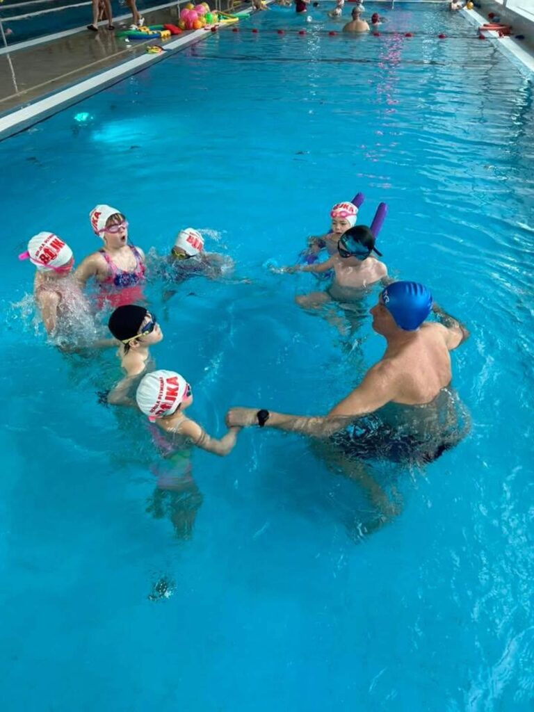 Na zdjęciu w krytym basenie Aqua Centrum Kolbudy odbywa się lekcja pływania. Siedmioro dzieci w czepkach i okularach stoi w kręgu razem z instruktorem. Trener w niebieskim czepku i kąpielówkach pomaga dwójce dzieci, pokazując ćwiczenie zespołowe. W tle widać innych pływających, a cała scena pokazuje radość, współpracę i zabawę w bezpiecznej wodzie.