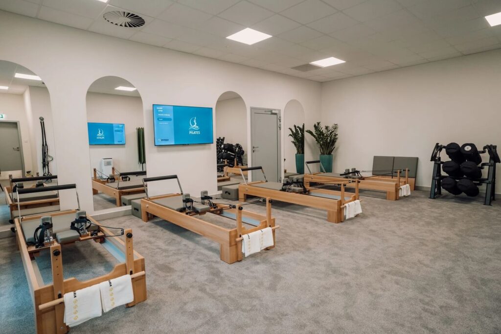 Na zdjęciu nowoczesne studio pilates urządzone w minimalistycznym stylu. Jasne wnętrze, białe ściany i równomierne oświetlenie tworzą spokojną atmosferę. Drewniane reformery ustawione są w równych rzędach na wykładzinie, każdy z nich wyposażony w ręcznik. Lustra po jednej stronie optycznie powiększają przestrzeń. Na ścianie widoczny ekran z napisem „PILATES”. W rogu sali znajdują się maty, czarne piłki do ćwiczeń i rośliny, które dodają wnętrzu przyjaznego charakteru.