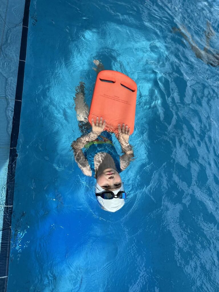 Na zdjęciu widać dziecko ćwiczącą w krytym basenie. Ma na sobie biały czepek i okularki zaciągnięte na czole. Trzyma oburącz pomarańczową deskę wypornościową przy krawędzi basenu, skupione na doskonaleniu techniki pływania.
