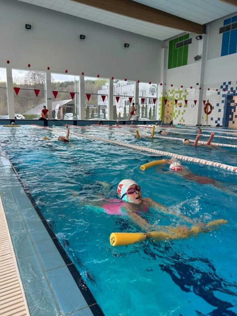 Na zdjęciu widzimy zorganizowaną lekcję pływania w krytym basenie. Kilkoro dzieci w czepkach i okularach pływackich ćwiczy w wodzie z pomocą żółtych makaronów piankowych, co wskazuje na trening techniczny lub zajęcia dla początkujących. Tory są wyraźnie oddzielone, a nad basenem wiszą czerwone chorągiewki — typowe oznaczenia dla stylu grzbietowego. Przestrzeń jest jasna, z dużymi oknami wpuszczającymi naturalne światło, kolorowymi panelami na ścianach i obecnością osób poza wodą.