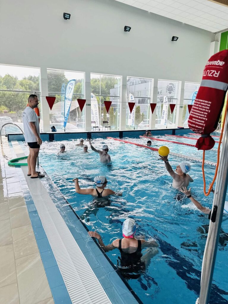 Na zdjęciu widzimy dynamiczną scenę z krytego basenu, gdzie trwa trening lub rozgrywka wodna — prawdopodobnie element zajęć z piłki wodnej. Kilkoro uczestników w czepkach znajduje się w wodzie, a jedna osoba unosi nad powierzchnię żółtą piłkę, przygotowując się do rzutu. Na brzegu stoi instruktor lub trener, uważnie obserwując przebieg ćwiczenia. Basen jest podzielony linami torowymi, a nad wodą wiszą czerwone chorągiewki, typowe dla oznaczeń w pływaniu. Przestrzeń jest jasna, z dużymi oknami i widokiem na zieleń.