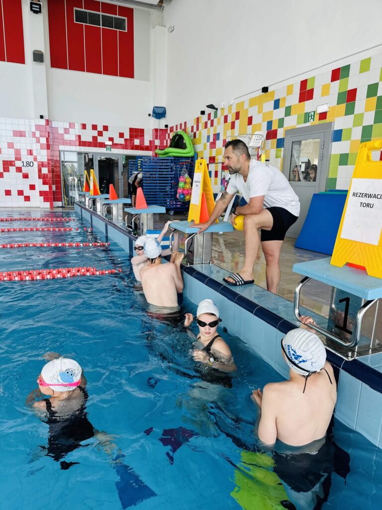 Na zdjęciu trening pływacki w krytym basenie. Kilkoro dzieci w czepkach i okularkach stoi w wodzie, skupionych wokół instruktora, który pokazuje ćwiczenie. Trener trzyma jedno z dzieci za rękę. W tle widać osoby korzystające z basenu, leżaki i zabawki przy brzegu. Woda jest czysta, a wnętrze jasne i uporządkowane.