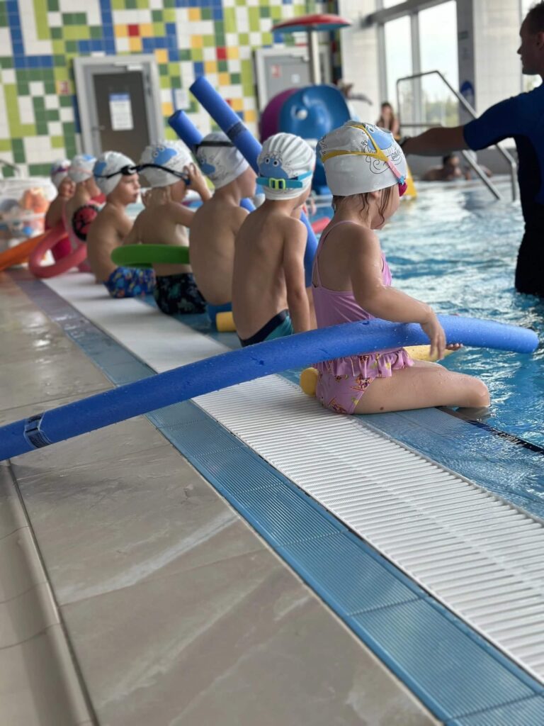 Na zdjęciu w krytym basenie siedzi grupa małych dzieci z niebieskimi makaronami piankowymi. Wszystkie mają czepki, stroje kąpielowe i patrzą na instruktora w wodzie, który pokazuje kolejne ćwiczenie. Instruktor ma na sobie czarny kombinezon. W tle widać kolorowe kafelki i duże okna wpuszczające światło.
