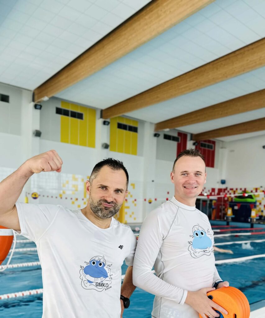 Zdjęcie przedstawia dwóch uśmiechniętych mężczyzn, znajdujących się w Aqua Centrum Kolbudy. Mężczyzna po lewej stronie ma na sobie białą koszulkę i jedną ręką pokazuje napięty biceps, drugi zaś ma założoną biała bluzkę i trzyma coś pomarańczowego w dłoniach. W tle widać basen z wyznaczonymi torami.