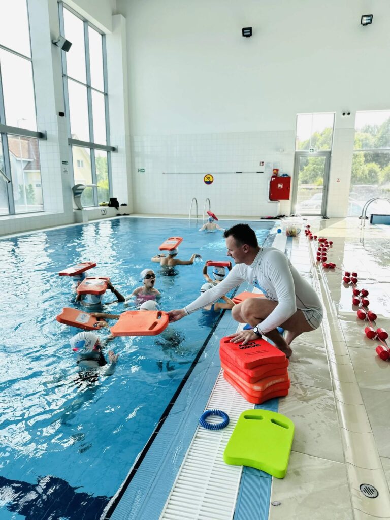 Na zdjęciu przedstawiono zorganizowaną lekcję pływania w jasnym, przestronnym obiekcie. Kilkoro dzieci znajduje się w wodzie, każde z nich trzyma pomarańczową deskę treningową, a instruktor przy brzegu rozdaje kolejne z przygotowanego stosu. Obok niego leży zielona deska, a wzdłuż krawędzi basenu ustawiono czerwono-białe hantle wypornościowe. Duże okna wpuszczają naturalne światło, a kolorowe kafelki i czysta przestrzeń podkreślają profesjonalny, bezpieczny charakter zajęć.