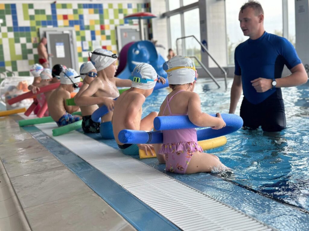 Na zdjęciu dzieci biorą udział w lekcji pływania w krytym basenie. Siedzą na brzegu w czepkach i strojach kąpielowych, trzymając kolorowe makarony piankowe. Instruktor w niebiesko-czarnym kombinezonie stoi w wodzie obok nich, gotowy do pomocy. W tle widać kolorowe kafelki, drzwi oraz sprzęt basenowy