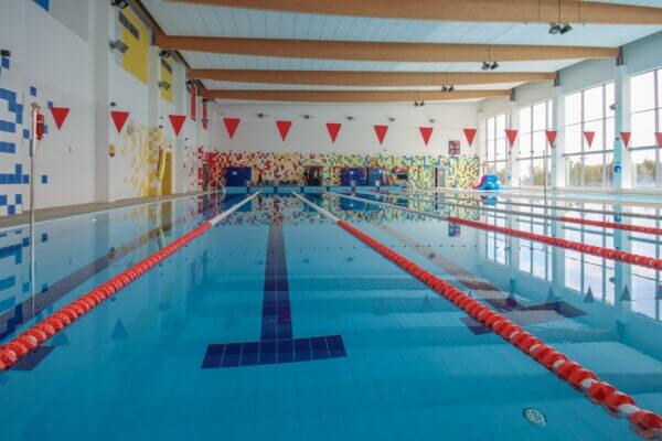 Na zdjęciu widzimy zadbany, kryty basen pływacki z kilkoma torami oddzielonymi czerwonymi linami. Woda jest przejrzysta i spokojna, a niebieskie płytki wyraźnie oznaczają linie torów oraz miejsca nawrotów. Nad basenem zawieszone są czerwone trójkątne chorągiewki, które służą jako wizualne punkty orientacyjne dla pływaków. Ściany ozdobiono kolorowymi kafelkami i wzorami, a duże okna po prawej stronie wpuszczają naturalne światło. Drewniane belki stropowe i lampy sufitowe dopełniają atmosfery nowoczesnego, przyjaznego obiektu sportowego.
