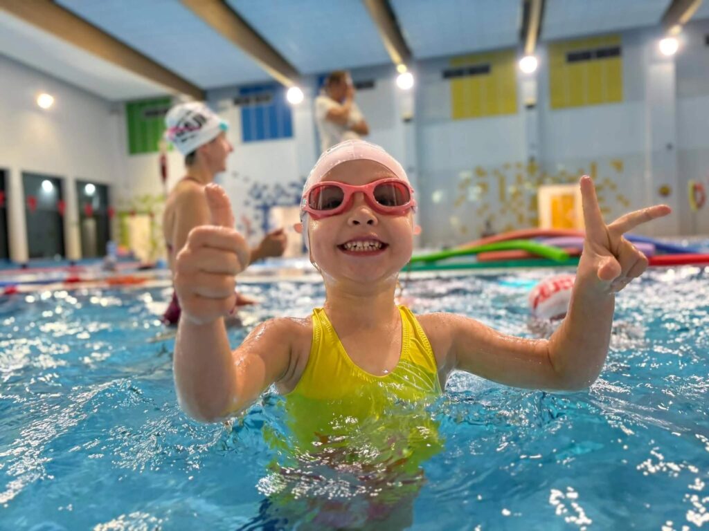 Na zdjęciu dziecko w żółtym stroju kąpielowym, różowym czepku i czerwonych okularach uśmiecha się i pokazuje „okej”. W tle są inne dzieci w wodzie i dorosły przy brzegu. Kolorowe panele i zabawki wodne tworzą przyjazną atmosferę.