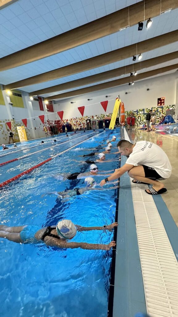 Na zdjęciu widać lekcję pływania w krytym basenie. Kilkoro dzieci w czepkach i okularach stoi przy brzegu, a instruktor w białej koszulce i czarnych spodenkach klęczy obok, tłumacząc ćwiczenie jednemu z uczestników. W tle widać czerwono-białe dekoracje na ścianach, cyfrowy zegar i drewniane belki sufitowe, które nadają sali nowoczesny charakter.