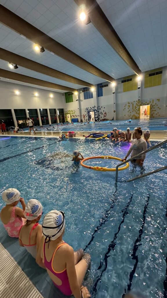 Na zdjęciu widzimy lekcję pływania w krytym basenie, prowadzoną w przyjaznej i kolorowej przestrzeni. Grupa dzieci w czepkach i strojach kąpielowych siedzi na brzegu basenu, uważnie obserwując instruktora, który znajduje się w wodzie i prowadzi ćwiczenia z wykorzystaniem kolorowych obręczy. W tle widać innych uczestników oraz osoby dorosłe.