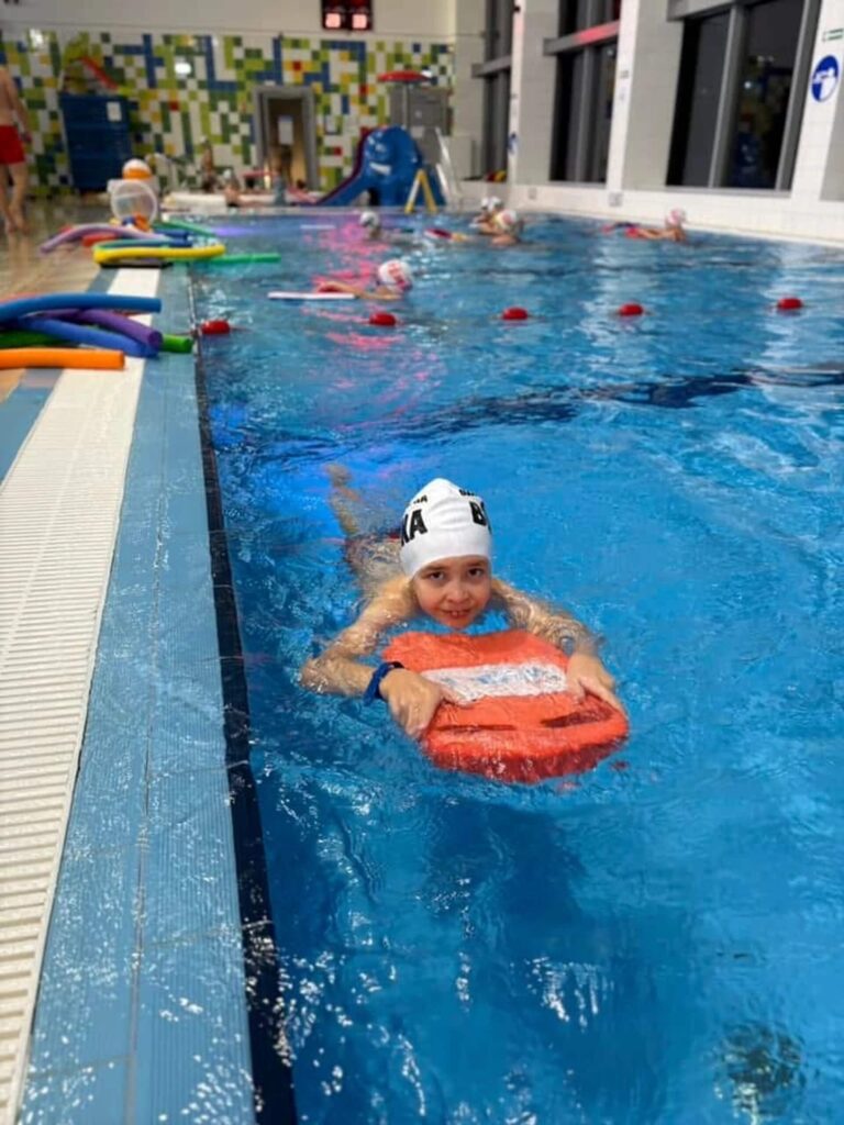 Zdjęcie przedstawia dziecięce zajęcia na krytym basenie. Na pierwszym planie młody pływak w białym czepku trzyma pomarańczową deskę do nauki pływania, unosząc się blisko brzegu. W tle widać inne dzieci uczestniczące w lekcji, a na brzegu rozłożony jest kolorowy sprzęt treningowy. Basen podzielony jest na tory, co nadaje przestrzeni uporządkowany, szkoleniowy charakter. Ściany wyłożone są kafelkami, a całość oświetlona jest miękkim światłem, tworząc atmosferę skupienia i bezpieczeństwa. Kompozycja emanuje ruchem, nauką i pozytywną energią — to scena zorganizowanych zajęć, które wspierają rozwój umiejętności i pewności siebie w wodzie.