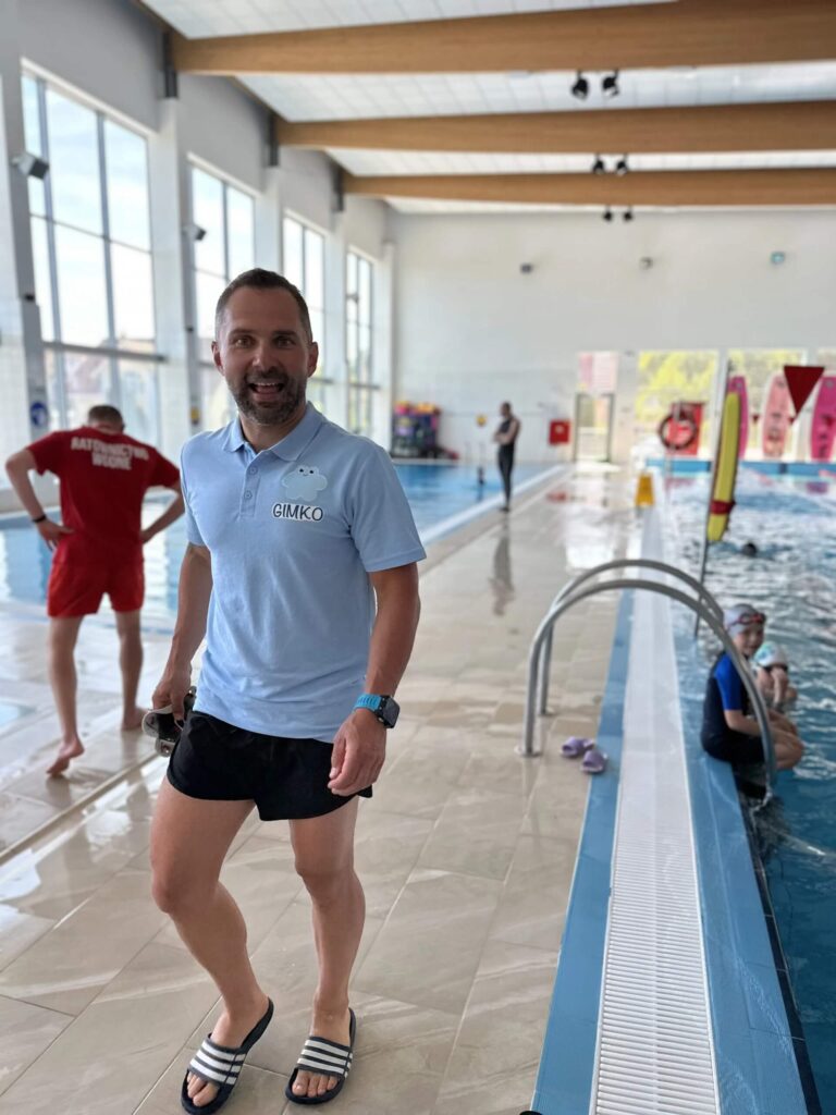 Zdjęcie przedstawia mężczyznę w jasnej koszulce z napisem ,,GIMKO'', który stoi przy brzegu basenu. W wodzie można zauważyć kilka osób. W tle widoczny jest ratownik ubrany w czerwony strój. Po lewej stronie zdjęcia widoczne duże okna, a na suficie drewniane belki.
