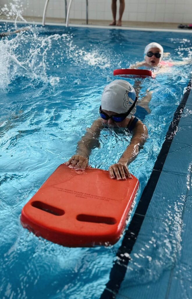 Na zdjęciu uchwycono moment treningu pływackiego w krytym basenie. Dwójka dzieci w czepkach i okularach pływackich płynie wzdłuż toru, korzystając z pomarańczowych desek wypornościowych. Widoczne rozpryski wody świadczą o intensywnej pracy nóg i dynamicznej aktywności.