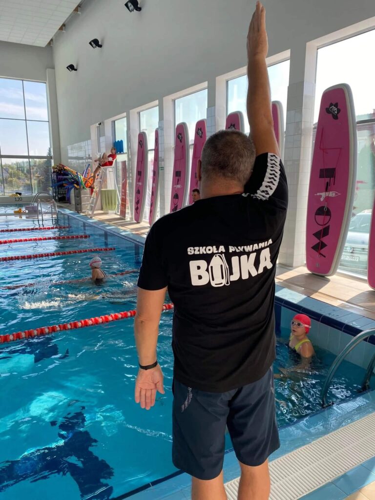 Zdjęcie przedstawia zajęcia nauki pływania w Aqua Centrum Kolbudy. Na brzegu widoczny instruktor w czarnej koszulce z napisem ,,Szkoła pływania BOJKA'' i granatowych spodenkach unosi jedną rękę do góry. Instruktor stoi tyłem do obiektywu. W wodzie znajdują się osoby w czepkach, a tory pływackie są wyraźnie oddzielone czerwonymi liniami. Po prawej stronie ustawiono pionowo różowe deski ratunkowe lub treningowe, co podkreśla szkoleniowy charakter zajęć. Całość odbywa się w jasnym, przestronnym wnętrzu z dużymi oknami.