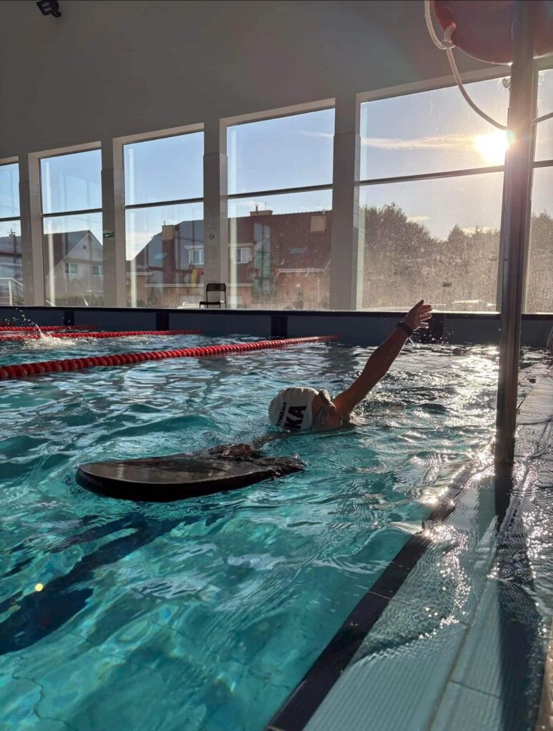 Na zdjęciu przedstawiona została osoba, która ćwiczy w krytym basenie Aqua centrum Kolbudy, płynąc z pomarańczową deską. Jedna ręka jest uniesiona nad wodą, druga spoczywa na desce. Przez duże okna w tle wpada światło zachodzącego słońca i odbija się na wodzie. Za oknami widać drzewa i budynki miasta.