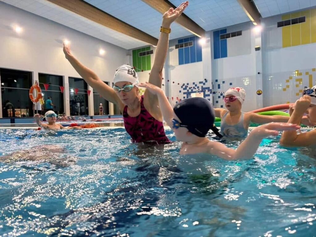 Na zdjęciu widać lekcję pływania w krytym basenie. Instruktorka w czepku i okularach pokazuje dzieciom ruchy ramion, a grupa maluchów uważnie ją naśladuje. Wszystkie dzieci mają czepki i okularki. W tle widać kolorowe kafelki, duże okna wpuszczające światło oraz sprzęt ratowniczy, w tym koła ratunkowe.