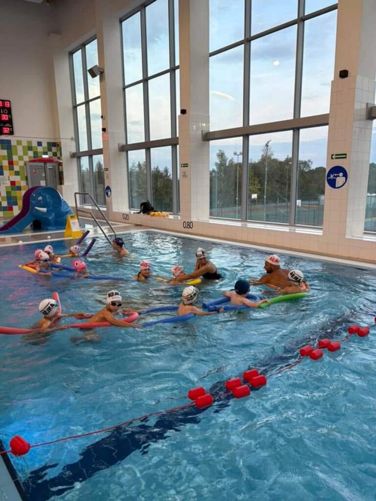 Na zdjęciu w krytym basenie Aqua Centrum Kolbudy widać lekcję pływania dla dzieci w płytkiej wodzie. Wszystkie uczestniczące dzieci mają na głowach czepki, bawią się kolorowymi makaronami piankowymi, unosząc się na wodzie w bezpieczny sposób. W tle znajduje się zjeżdżalnia. W oddali na ścianie widoczne kolorowe kafelki, a po prawej stronie duże okna wpuszczające naturalne światło.