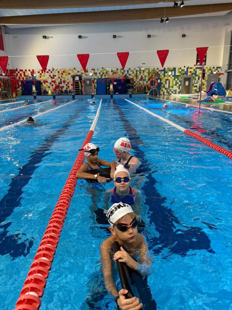 Na zdjęciu dzieci stoją w wodzie przy torze pływackim w krytym basenie Aqua Centrum Kolbudy. Mają na sobie czepki i okularki, ustawione są wzdłuż liny torowej, przygotowane do zajęć. W tle widać kolorowe kafelki na ścianie oraz czerwone chorągiewki zawieszone nad końcem toru. W oddali można zauważyć niebieską zjeżdżalnię.