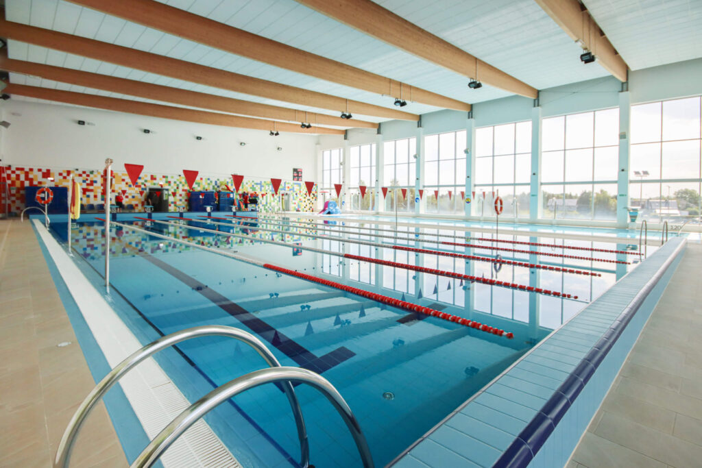 Zdjęcie przedstawia nowoczesny kryty basen z kilkoma torami oddzielonymi czerwonymi linami, co sugeruje przestrzeń przeznaczoną do pływania sportowego lub treningowego. Przez duże okna sięgające od podłogi do sufitu wpada naturalne światło, rozświetlając wnętrze i podkreślając czystość oraz przestronność obiektu. Drewniane belki stropowe i wiszące lampy nadają przestrzeni ciepły, architektonicznie dopracowany charakter. Na dalszej ścianie widoczna jest mozaika w odcieniach czerwieni, pomarańczu i żółci, która dodaje energii i wizualnego akcentu. Całość emanuje profesjonalizmem, dynamiką i spokojem, tworząc idealne warunki do treningu, rekreacji lub zawodów.