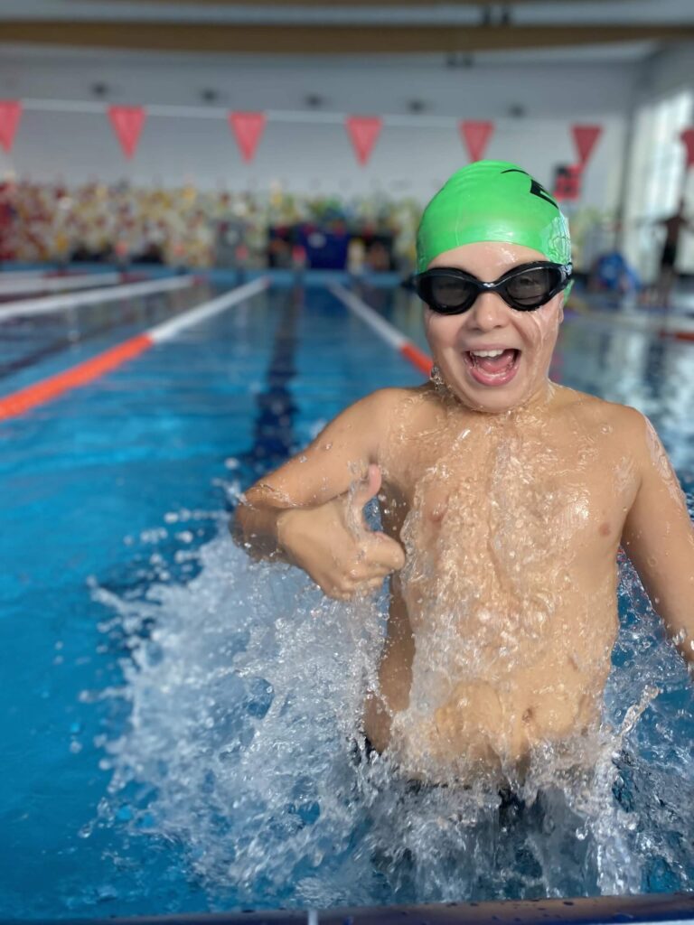 Na zdjęciu uchwycono dynamiczny moment — dziecko wynurza się z basenu, z szeroko otwartą buzią i uniesionym kciukiem w geście radości lub triumfu. Ma na sobie zielony czepek i czarne okularki, a wokół niego rozpryskuje się woda, podkreślając ruch i emocje chwili. W tle widać pomarańczowe linie torów, czerwone chorągiewki nad wodą oraz kolorowy mural na ścianie.