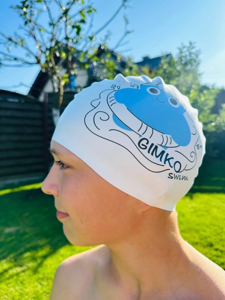 Na zdjęciu widać dziecko na świeżym powietrzu, ubrane w biały czepek pływacki z nadrukiem uśmiechniętej, niebieskiej rybki rozdymki otoczonej falami. Pod obrazkiem znajduje się napis „GIMKO SWIM”, prawdopodobnie oznaczający nazwę szkoły pływania, drużyny lub marki sportowej. Tło jest zamazane, ale można dostrzec zieloną trawę, drzewo i ciemne ogrodzenie.