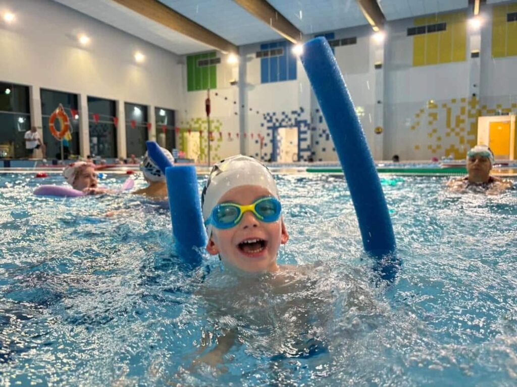 Na zdjęciu w krytym basenie widać dziecko w czepku i okularach, które bawi się w wodzie, trzymając niebieski makaron piankowy. Uśmiechnięte i swobodne, bierze udział w lekcji pływania. W tle widać inne dzieci korzystające z podobnych przyrządów do nauki i zabawy w wodzie.