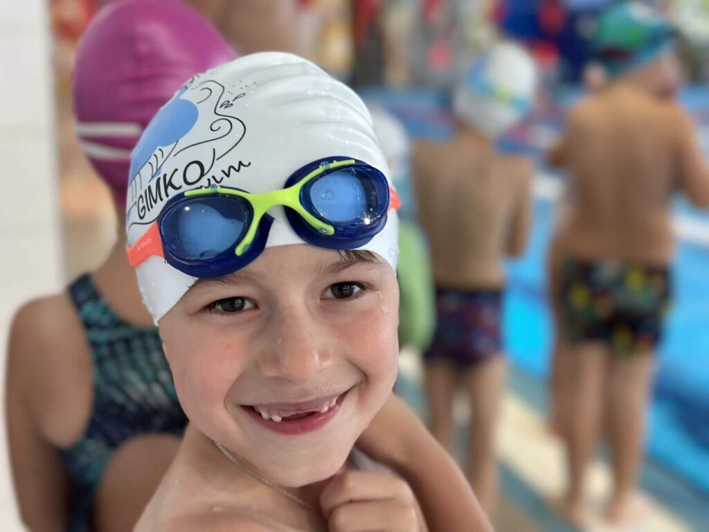 Na zdjęciu widzimy uśmiechnięte dziecko w białym czepku pływackim, na którym widnieje niebieski nadruk z napisem „GIMKO” i dynamicznym motywem wodnego rozbryzgu. Na czepku spoczywają zielono-niebieskie okularki, a w tle widać innych uczestników zajęć — również w czepkach i strojach kąpielowych, stojących przy basenie.