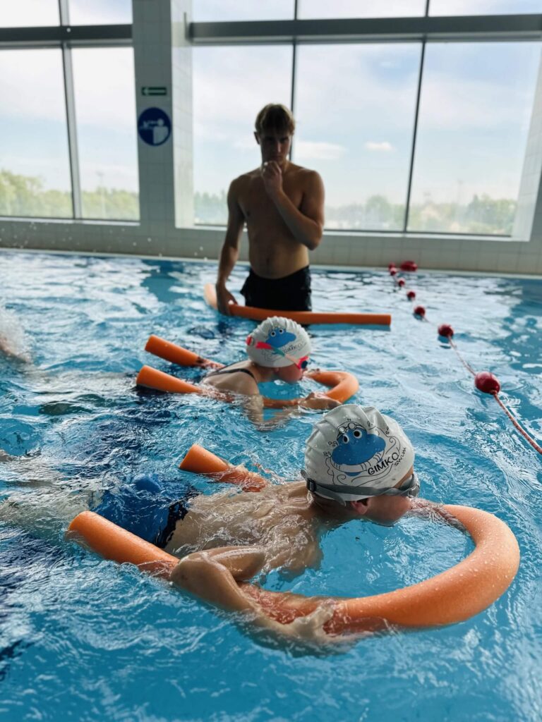 Na zdjęciu przedstawiona została lekcja pływania w krytym basenie. Dwoje dzieci w czepkach i okularach stoi w wodzie z pomarańczowymi makaronami. Instruktor w czarnych spodenkach stoi w wodzie przy krawędzi basenu, trzymając również pomarańczowy makaron. W tle widoczne są duże okna, które wpuszczają dużo światła.
