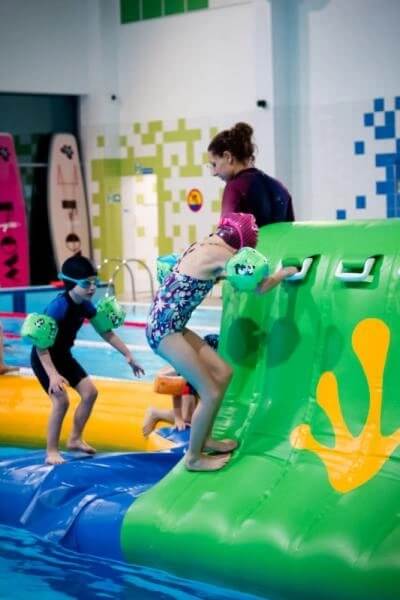 Na zdjęciu w krytym basenie grupa dzieci bierze udział w wodnej zabawie lub lekcji pływania. Jedno z dzieci wspina się po zielonej, dmuchanej konstrukcji z uchwytami, a inne stoi obok na niebieskiej powierzchni, mając na sobie czepek i zielone rękawki. W tle widać instruktora pomagającego wspinającemu się dziecku oraz kolorowe ściany i ustawione pionowo deski basenowe.