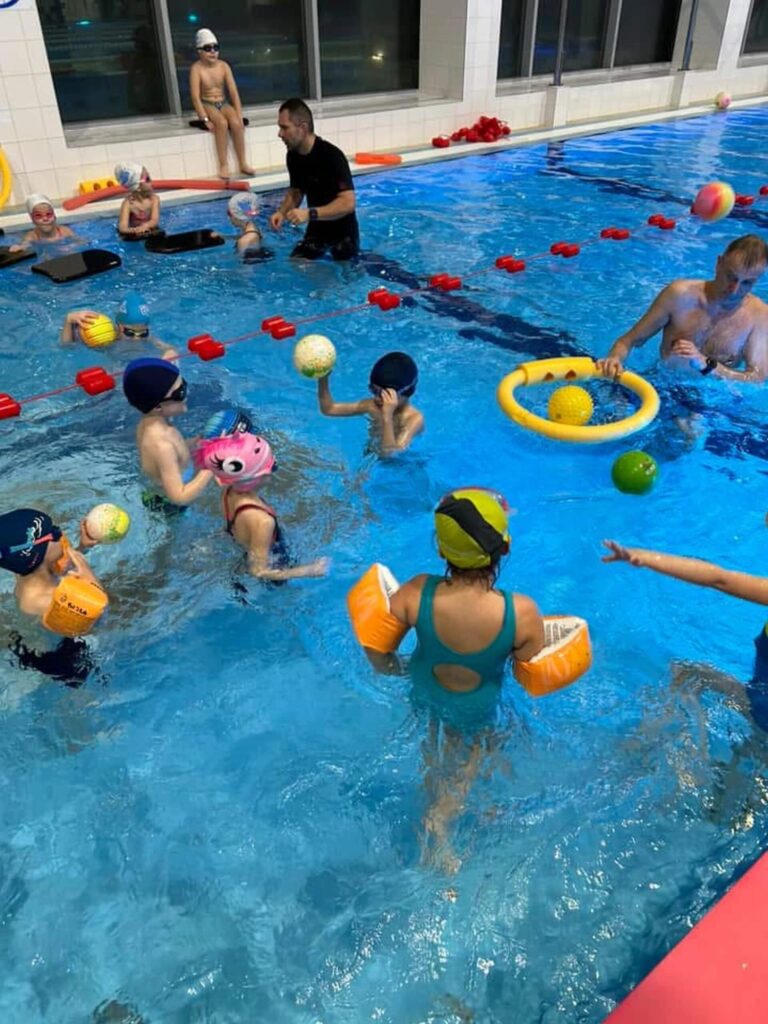 Na zdjęciu dzieci biorą udział w zajęciach na krytym basenie. Ubrane w czepki i rękawki, bawią się w wodzie, niektóre trzymają kolorowe piłki. Wśród nich znajduje się opiekun, który aktywnie uczestniczy w ćwiczeniach, drugi dorosły stoi przy brzegu. W basenie pływa żółte koło z piłkami, a czerwone linie wyznaczają tory. Jasne wnętrze i porządek wokół wskazują na dobrze zorganizowane zajęcia.